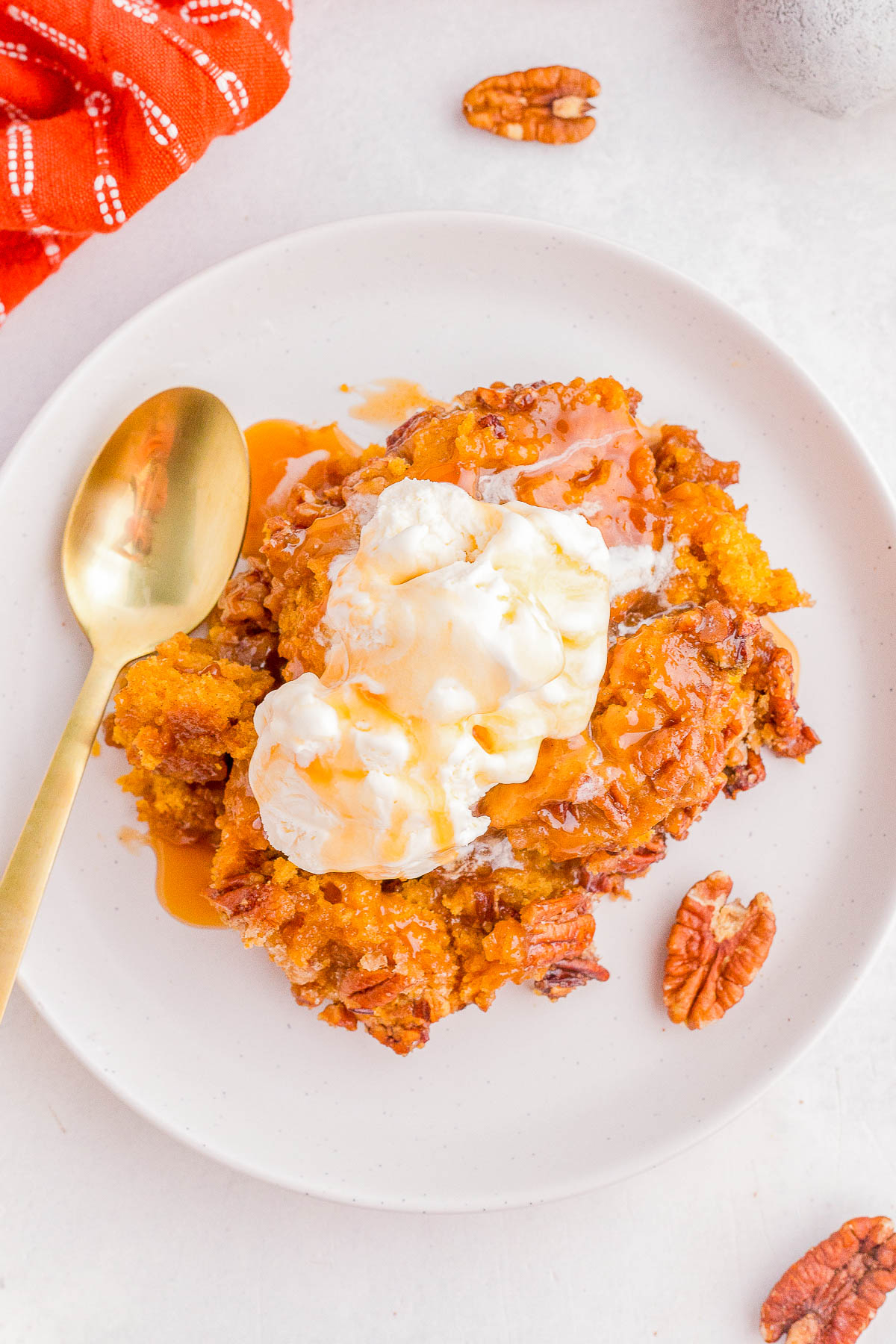 A plate with a serving of pumpkin dessert topped with vanilla ice cream and caramel sauce, next to a gold spoon and pecan halves on a white surface.