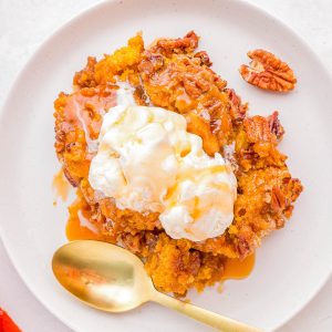 A serving of pumpkin dessert topped with a scoop of vanilla ice cream and drizzled with caramel sauce, garnished with pecans, on a white plate with a gold spoon.