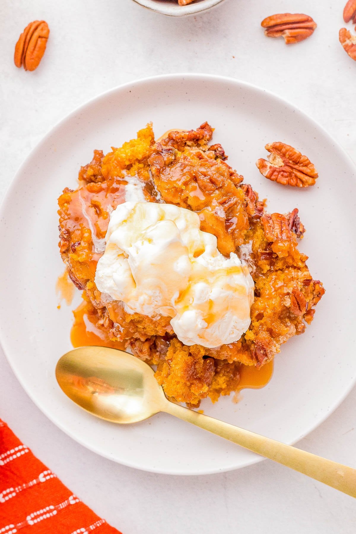 A serving of pumpkin dessert topped with a scoop of vanilla ice cream and drizzled with caramel sauce, garnished with pecans, on a white plate with a gold spoon.