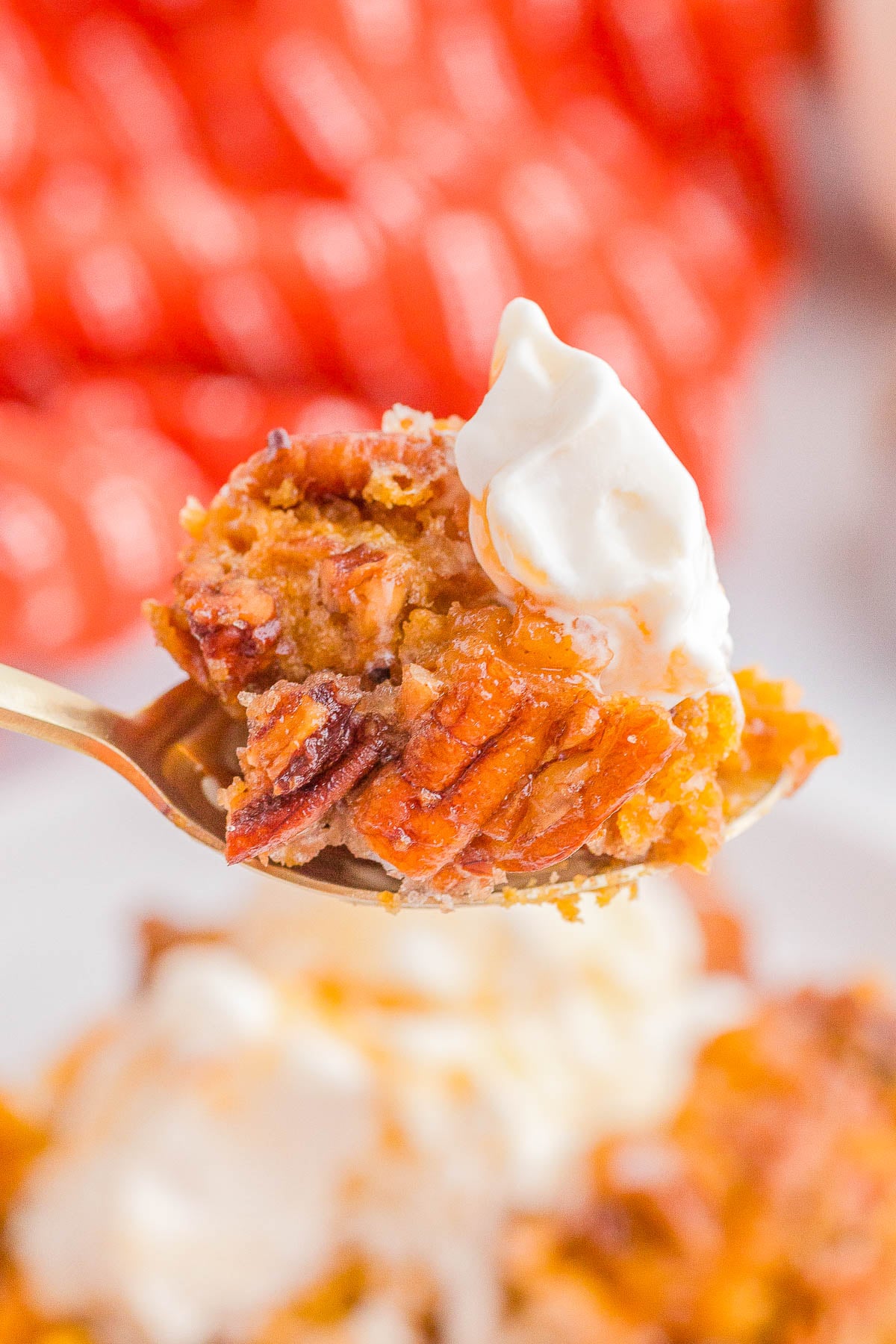 A close-up of a spoonful of pecan-topped dessert with a dollop of whipped cream, held in front of a blurred background.