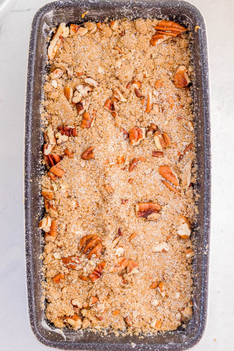 A rectangular loaf pan filled with unbaked crumb topping and chopped pecans on top, ready for baking.