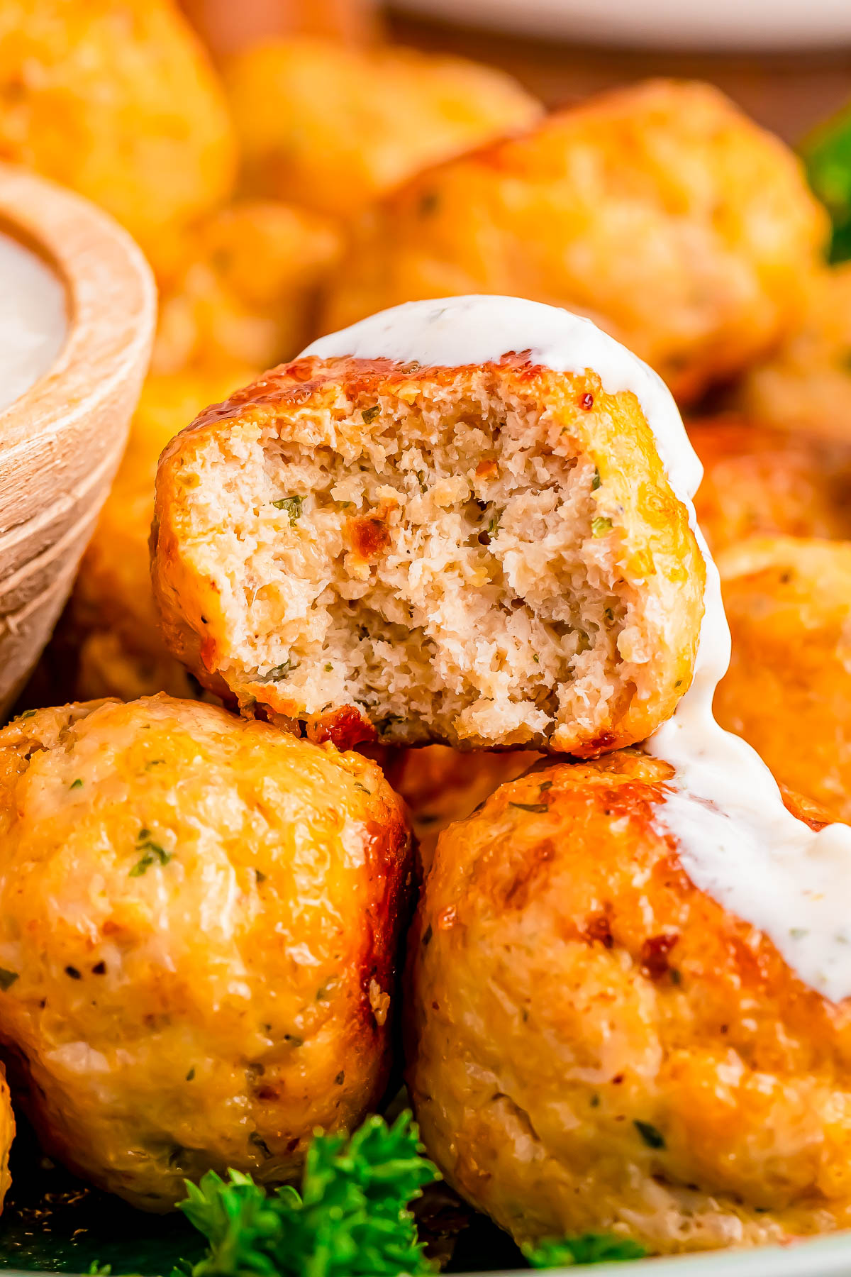Close-up of baked chicken meatballs, one with a bite taken out, topped with a drizzle of white sauce and garnished with fresh parsley.