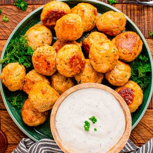 A plate of baked cheese balls with herbs is served with a bowl of creamy dipping sauce, garnished with parsley, on a wooden table with a striped napkin and fork.