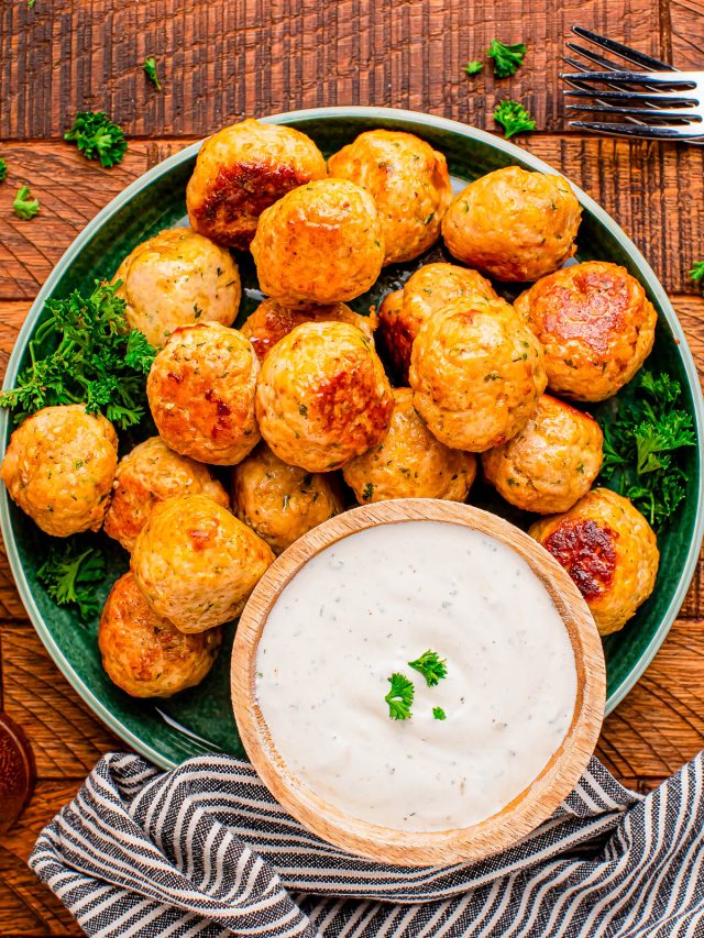 A plate of baked cheese balls with herbs is served with a bowl of creamy dipping sauce, garnished with parsley, on a wooden table with a striped napkin and fork.