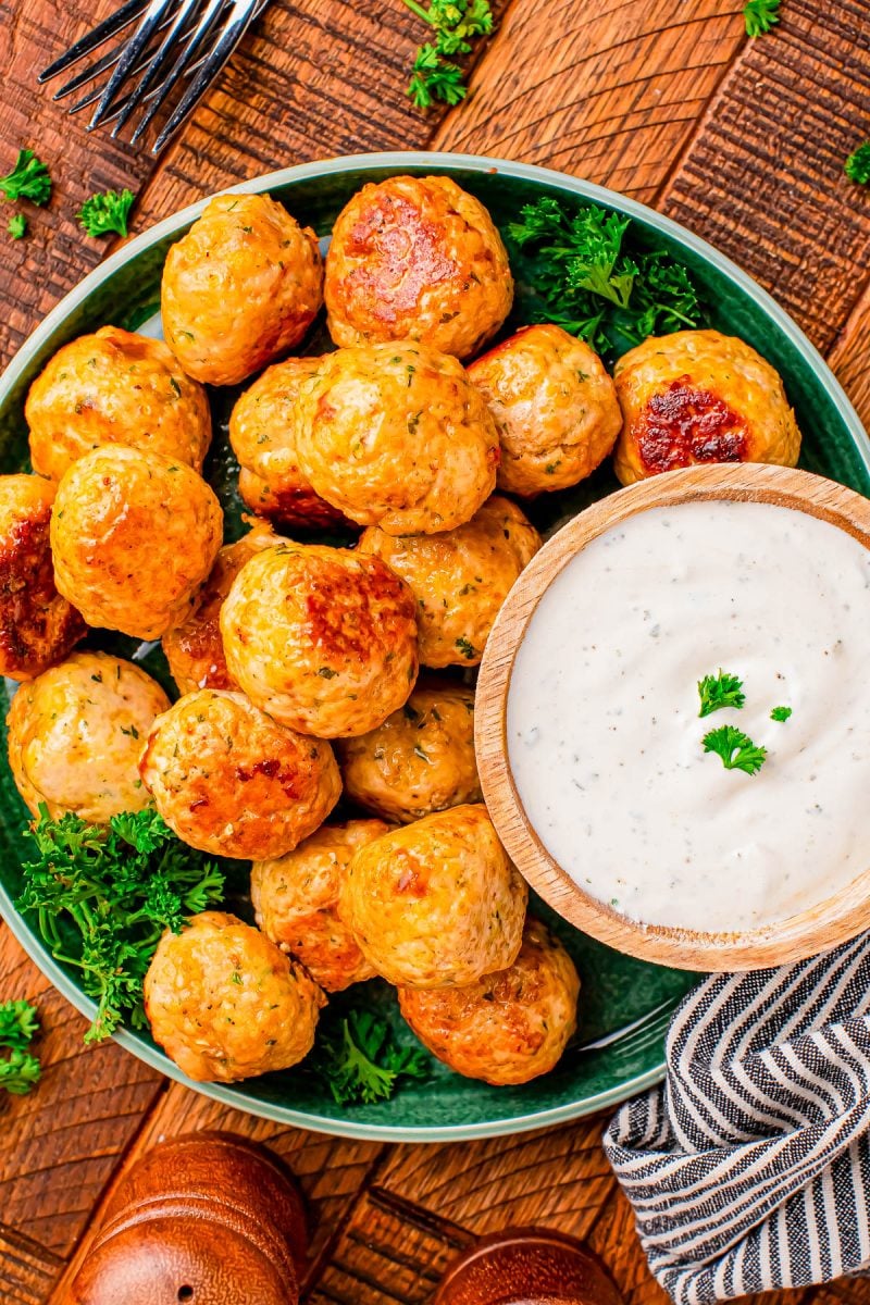 A plate of baked meatballs garnished with parsley, served with a bowl of creamy white dipping sauce on a wooden table.