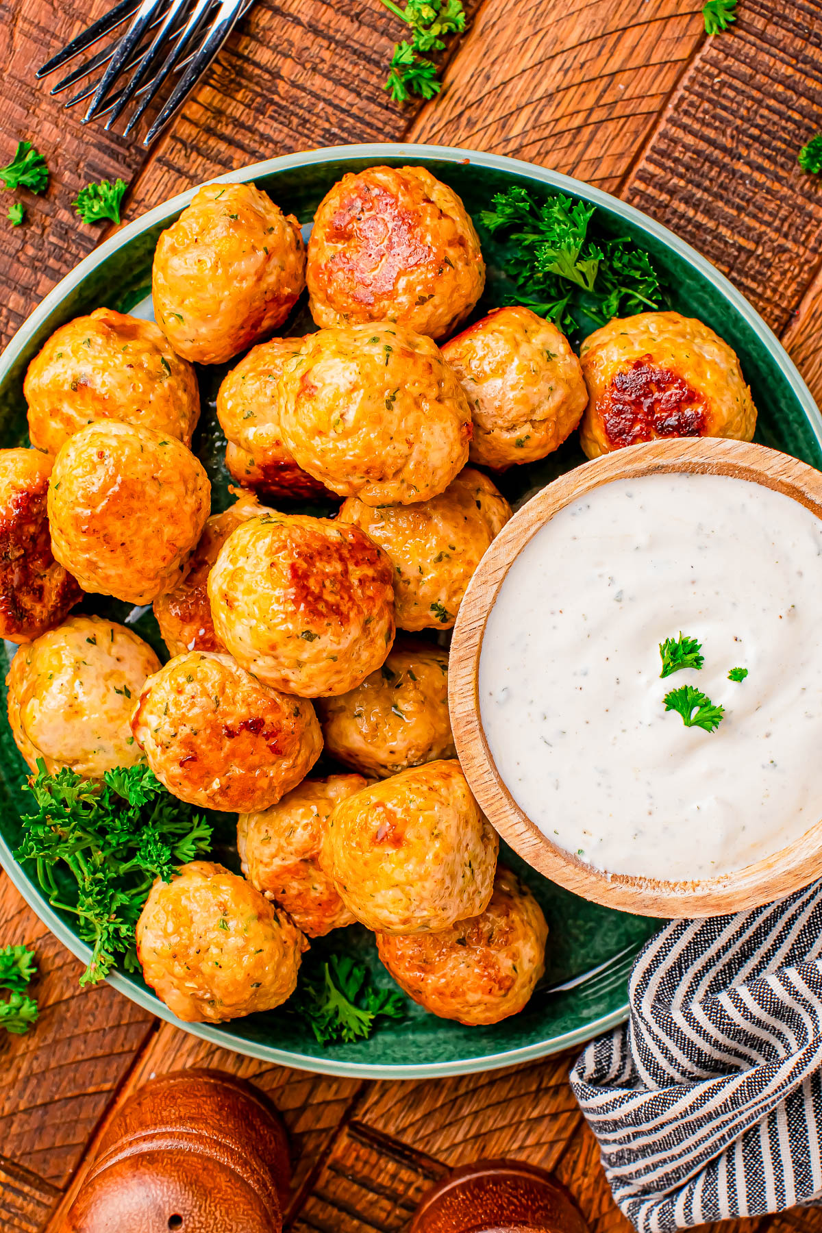 A plate of baked meatballs garnished with parsley, served with a bowl of creamy white dipping sauce on a wooden table.