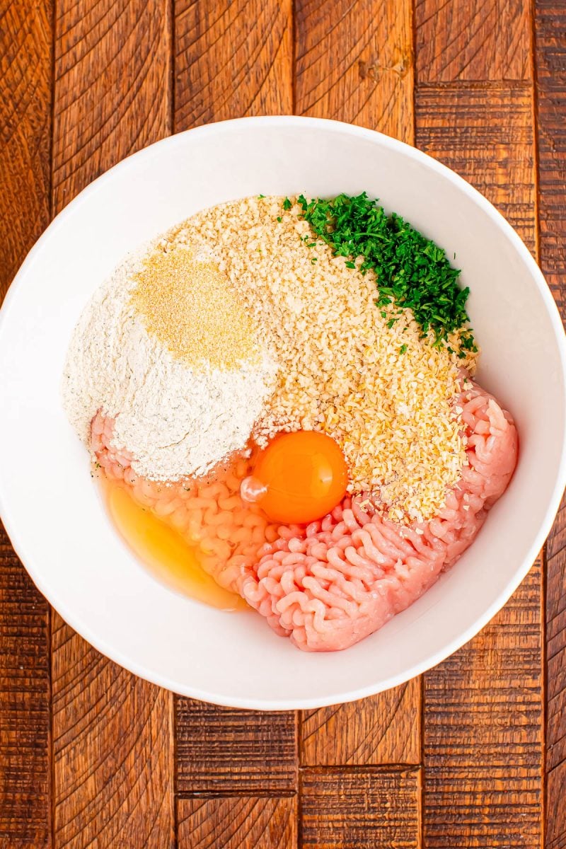 A white bowl containing ground meat, an egg, breadcrumbs, flour, dried seasoning, and chopped parsley on a wooden surface.