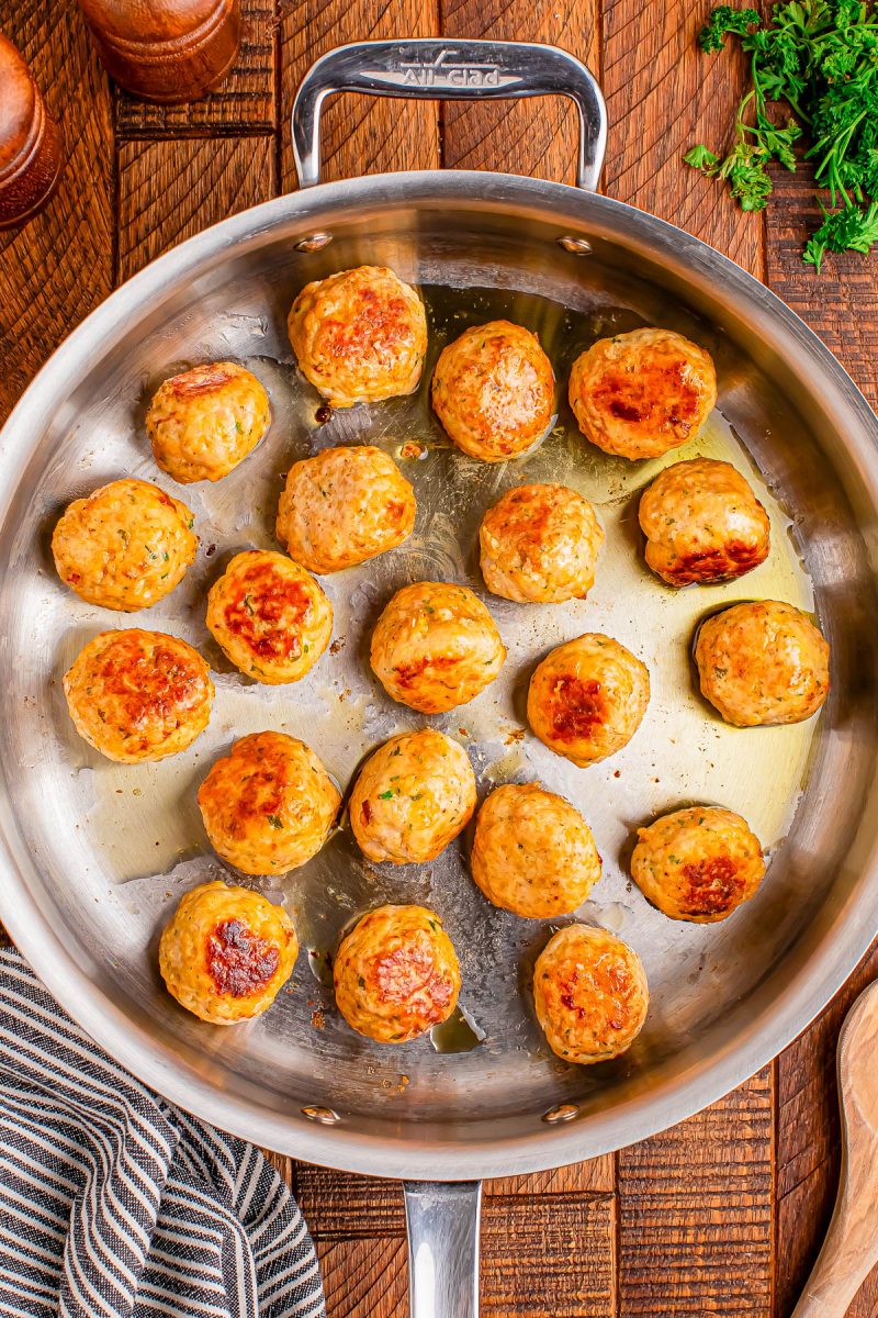 A stainless steel skillet with browned meatballs arranged inside, placed on a wooden surface with a striped towel and fresh parsley nearby.