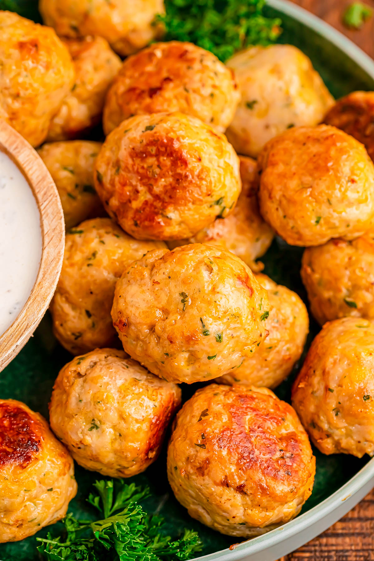 A plate of golden-brown baked meatballs garnished with parsley, served with a side of white dipping sauce in a small wooden bowl.