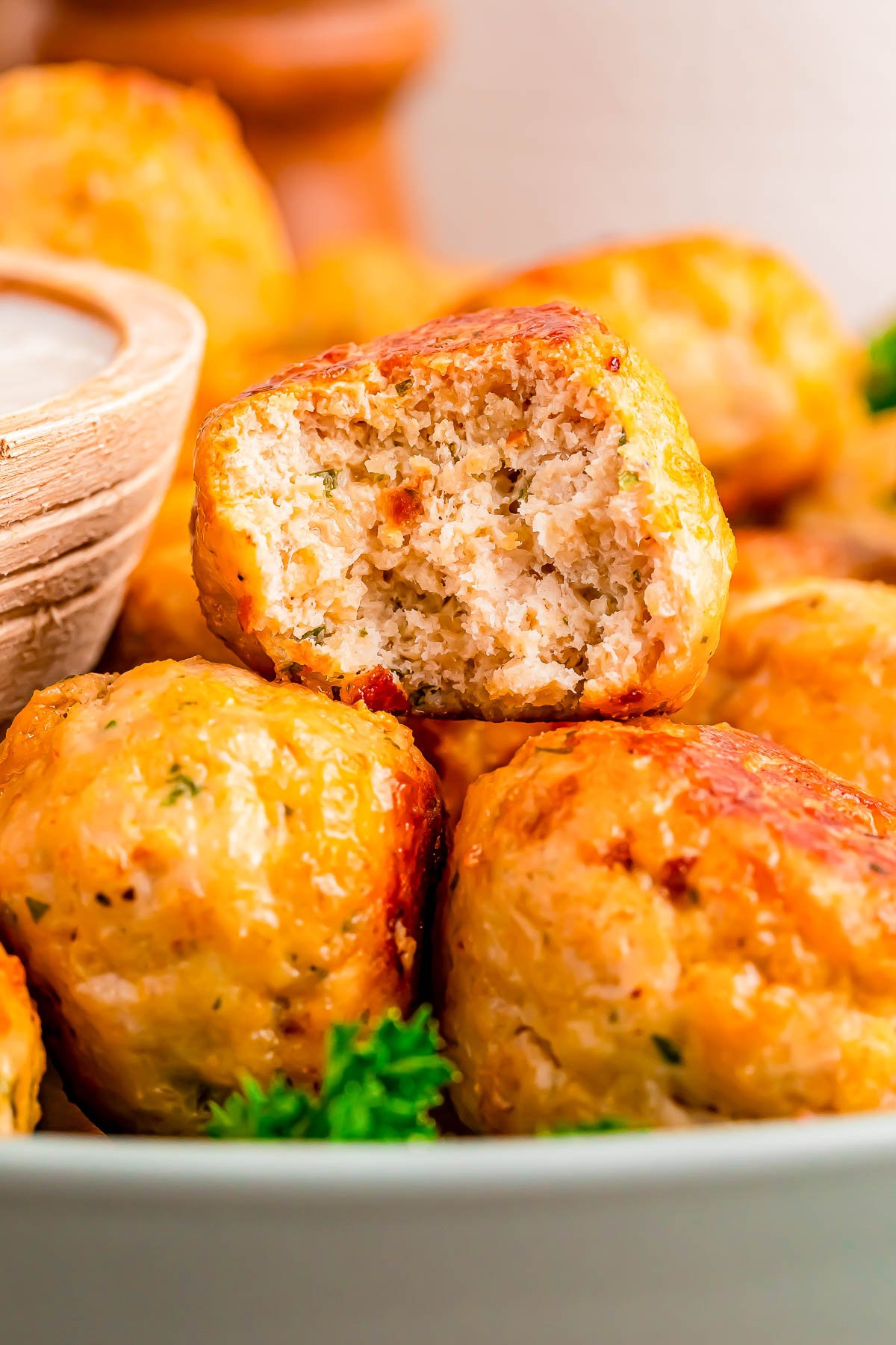A close-up of several baked chicken meatballs, one with a bite taken out, arranged in a bowl with a small wooden bowl of dipping sauce nearby.