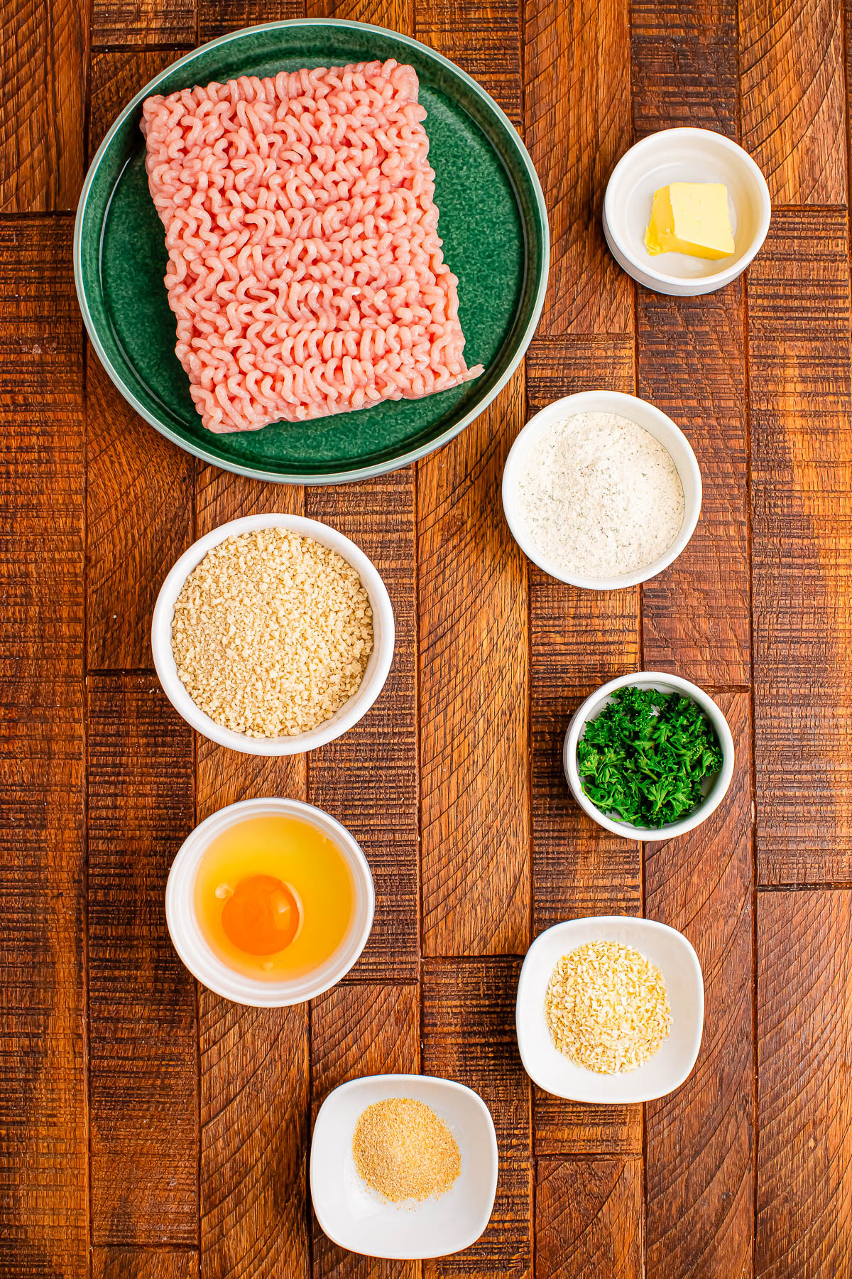 A plate of raw ground meat, with small bowls of butter, flour, breadcrumbs, chopped parsley, an egg, grated cheese, and garlic powder on a wooden surface.