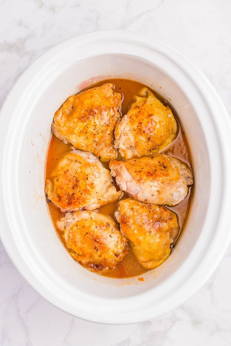 Six cooked chicken thighs with a golden brown surface arranged in a white slow cooker with visible juices at the bottom.