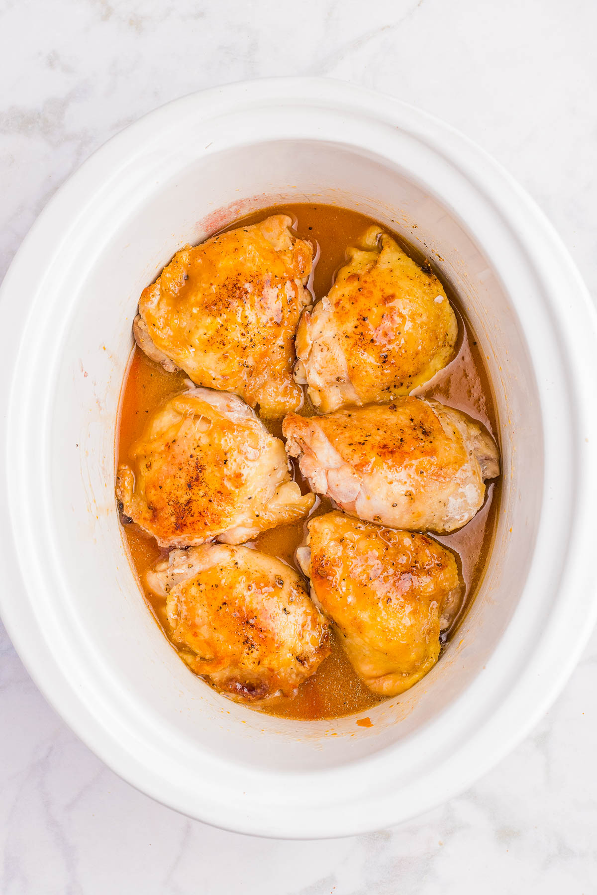 Six cooked chicken thighs with a golden brown surface arranged in a white slow cooker with visible juices at the bottom.