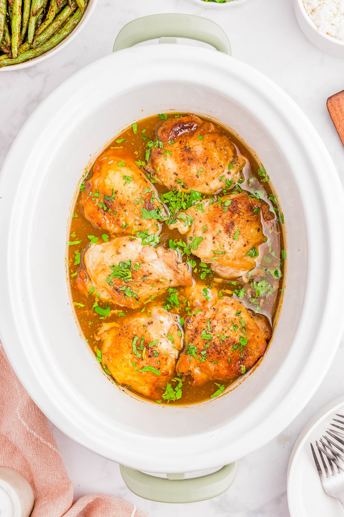 Five cooked chicken thighs in a slow cooker with a golden sauce, garnished with chopped parsley. Surrounding dishes include green beans and rice.