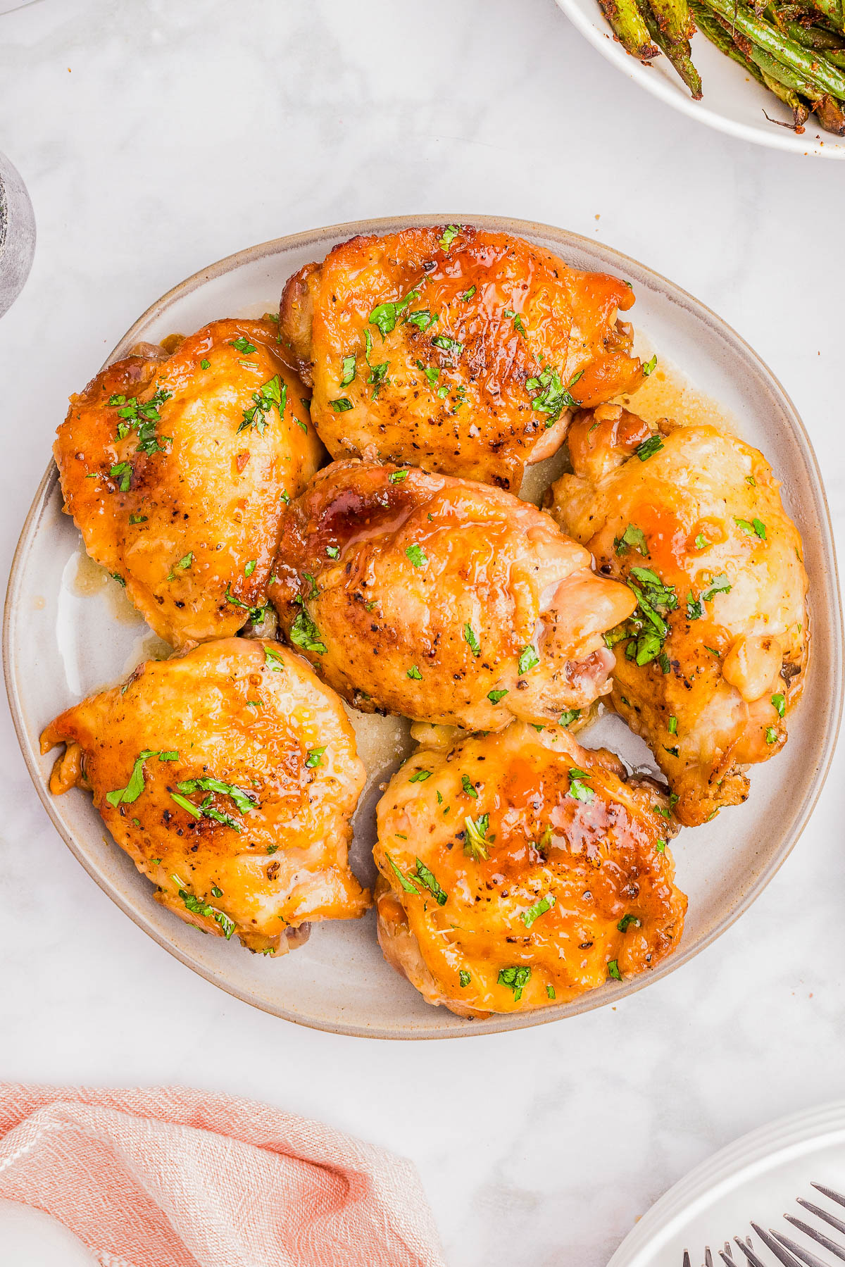 A plate with six cooked chicken thighs garnished with chopped herbs, placed on a white surface with a pink napkin nearby.