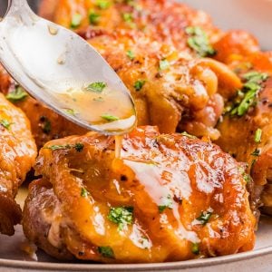 A spoon drizzles sauce over a piece of cooked, golden-brown chicken garnished with chopped herbs on a plate.