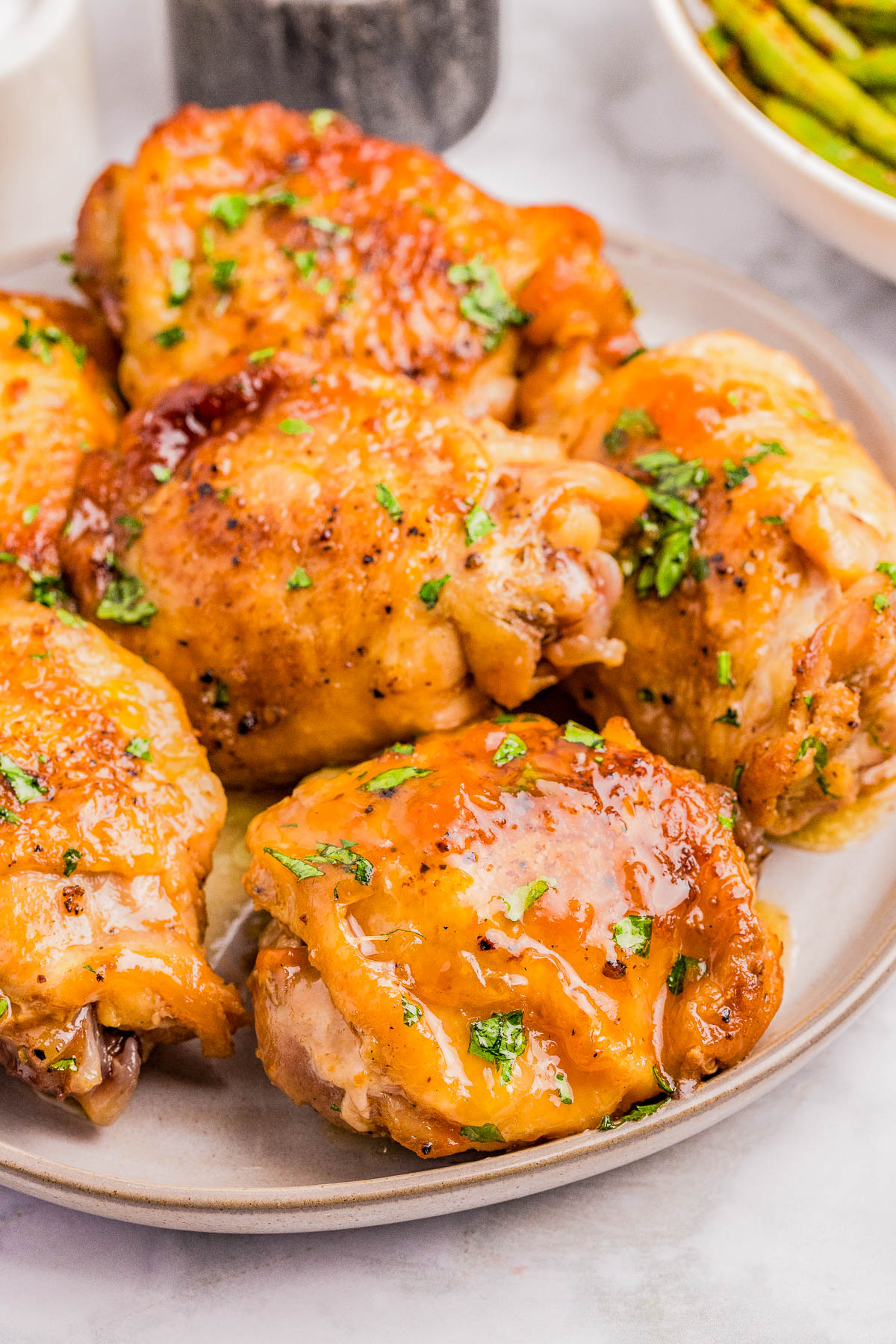Plate of glazed, baked chicken thighs garnished with chopped herbs, with a side of green beans partially visible in the background.