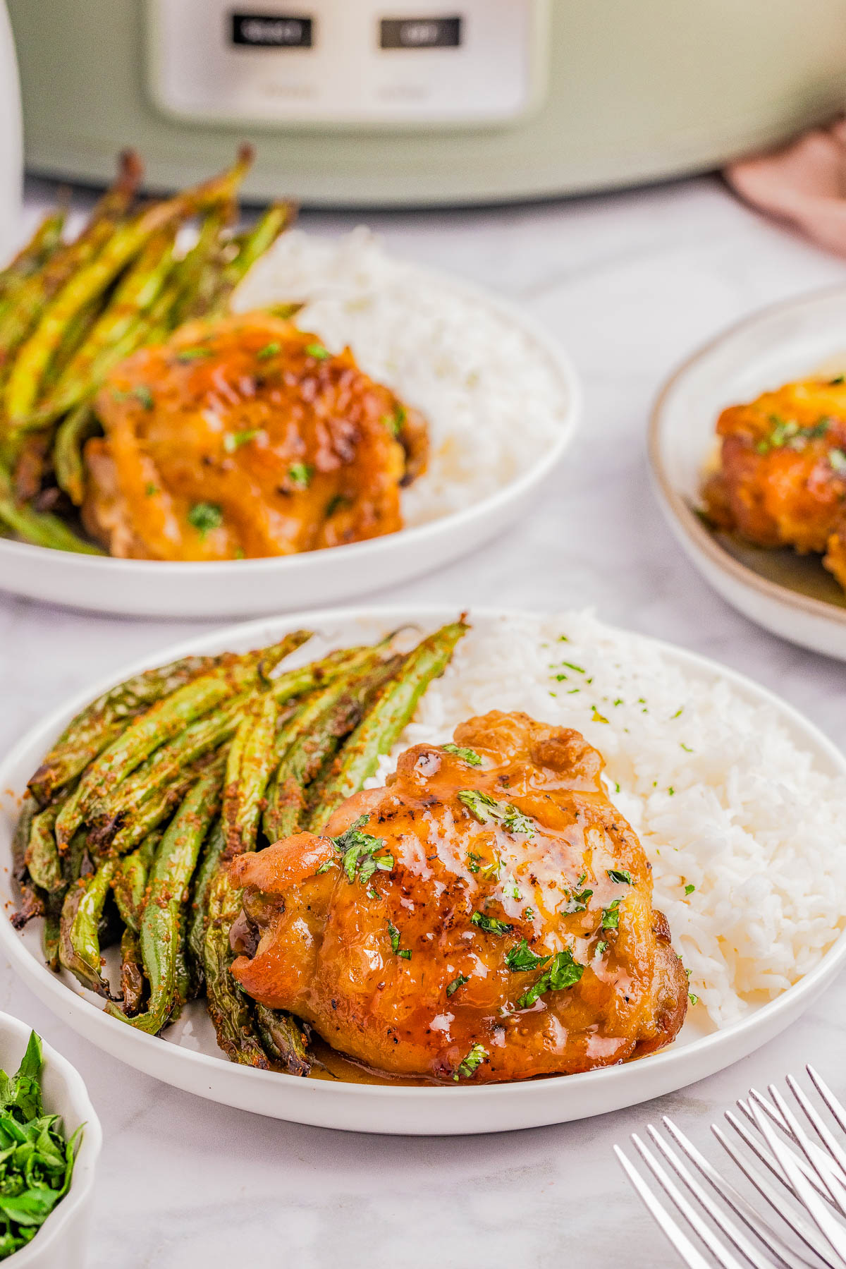 A plate of baked chicken thighs, white rice, and roasted green beans, with a fork and knife on the table and another similar plate in the background.