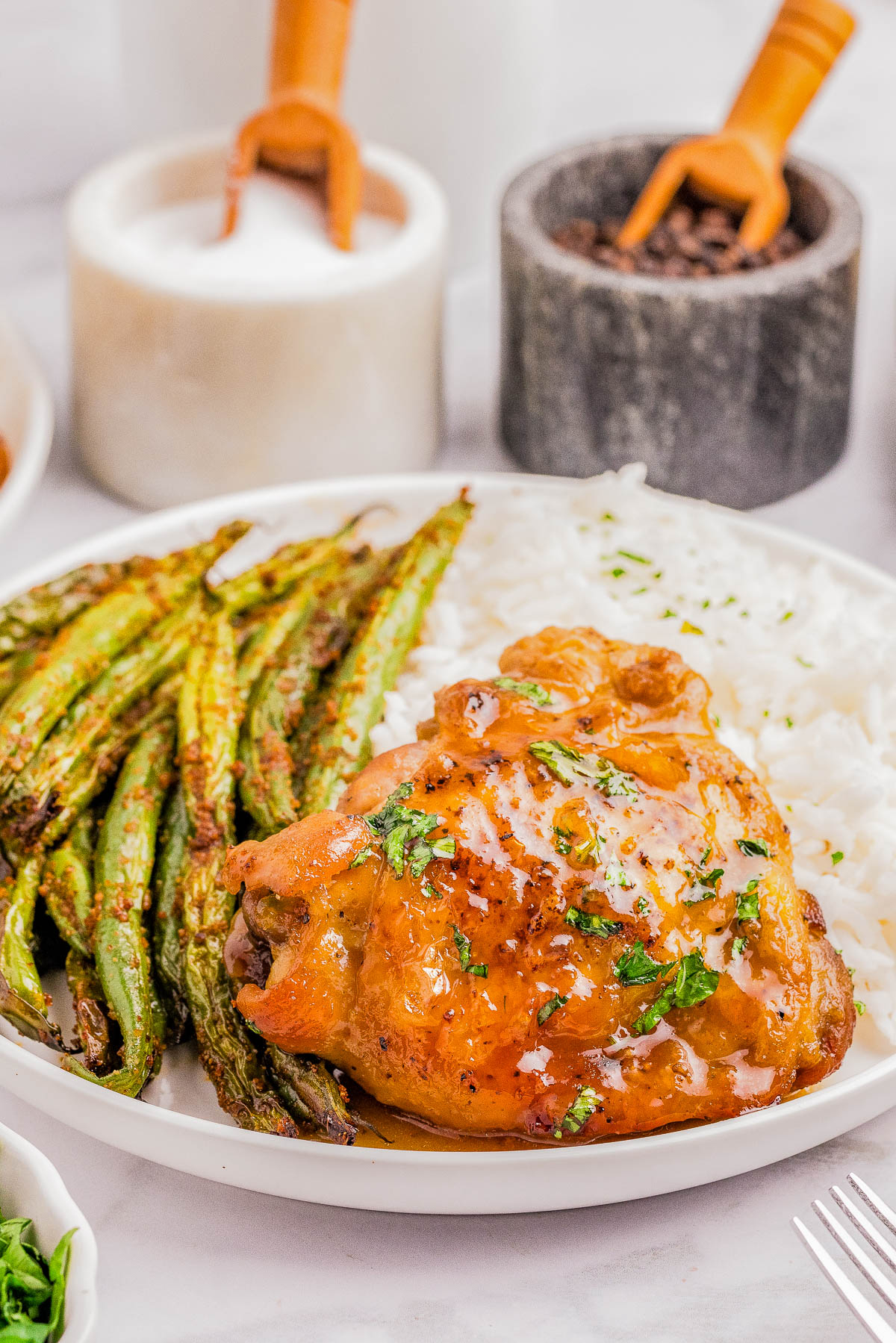A plate of glazed baked chicken, roasted green beans, and white rice, garnished with herbs. Salt and pepper containers with wooden spoons are in the background.