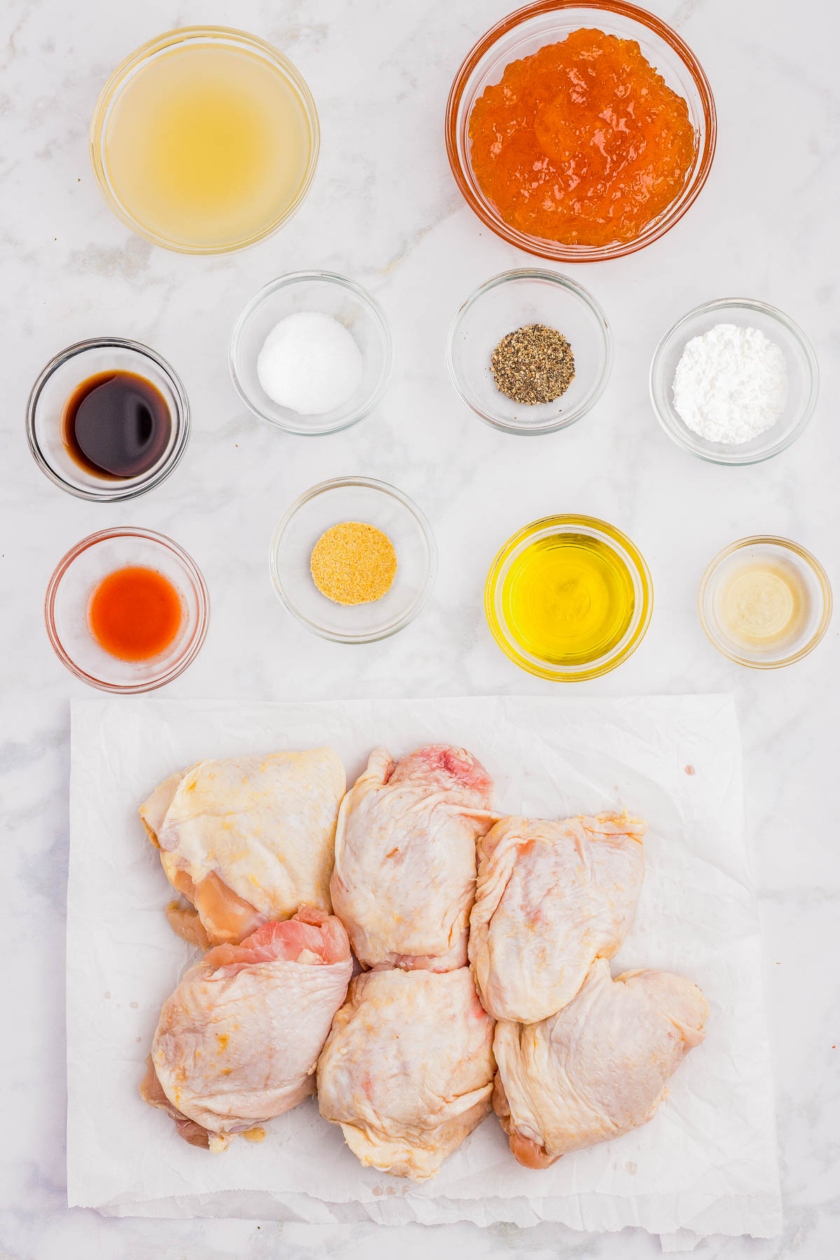 Raw chicken thighs on parchment paper are surrounded by small bowls containing various ingredients such as apricot preserves, seasonings, oil, vinegar, broth, and sauces on a white surface.