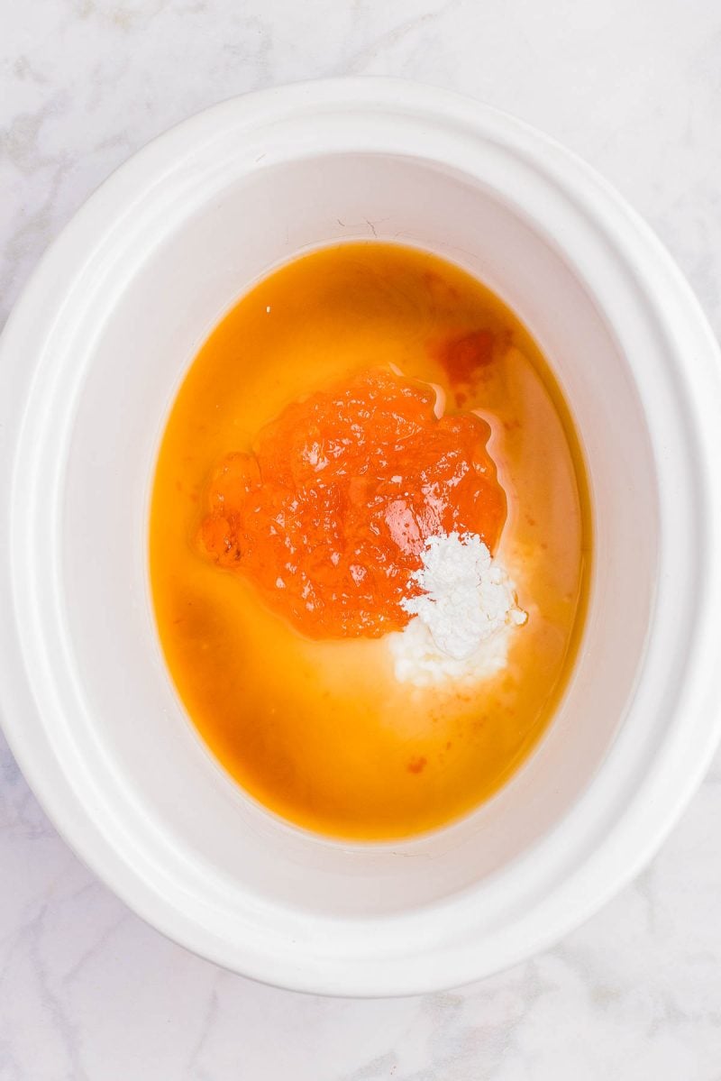White bowl containing orange-colored sauce, likely apricot preserves, with a white powder and liquid ingredients on a marble surface.