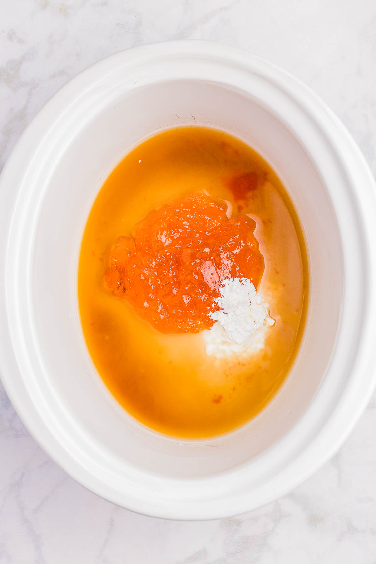 White bowl containing orange-colored sauce, likely apricot preserves, with a white powder and liquid ingredients on a marble surface.
