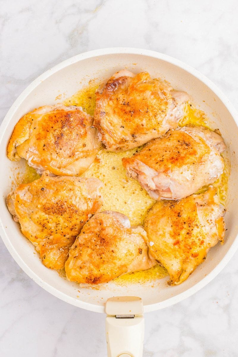 Six browned chicken thighs cooking in a white skillet with oil and seasonings on a marble countertop.