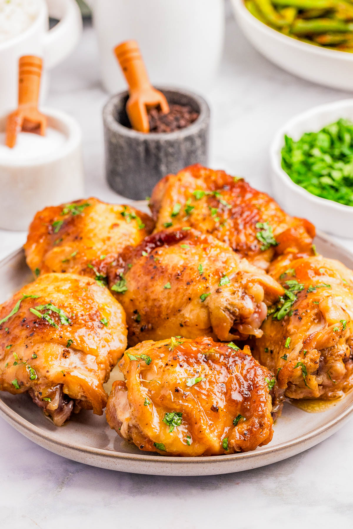 A plate of baked chicken thighs garnished with chopped herbs, with condiments and additional herbs in small bowls in the background.