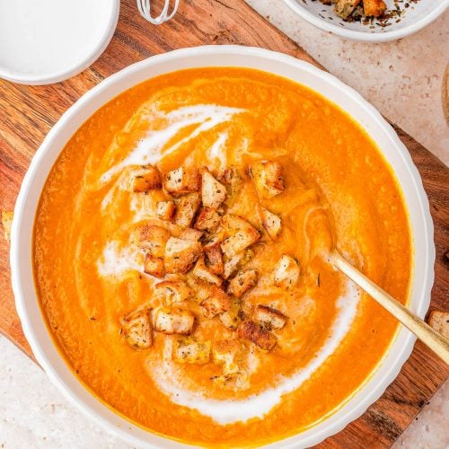 A bowl of creamy orange soup topped with golden-brown croutons and a swirl of cream, placed on a wooden board with extra croutons in a nearby bowl.