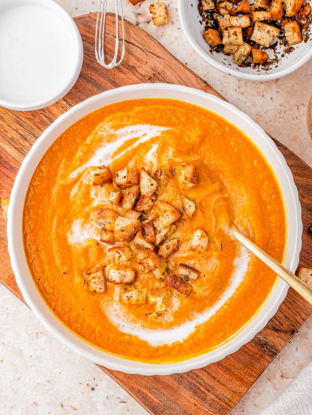 A bowl of creamy orange soup topped with golden-brown croutons and a swirl of cream, placed on a wooden board with extra croutons in a nearby bowl.