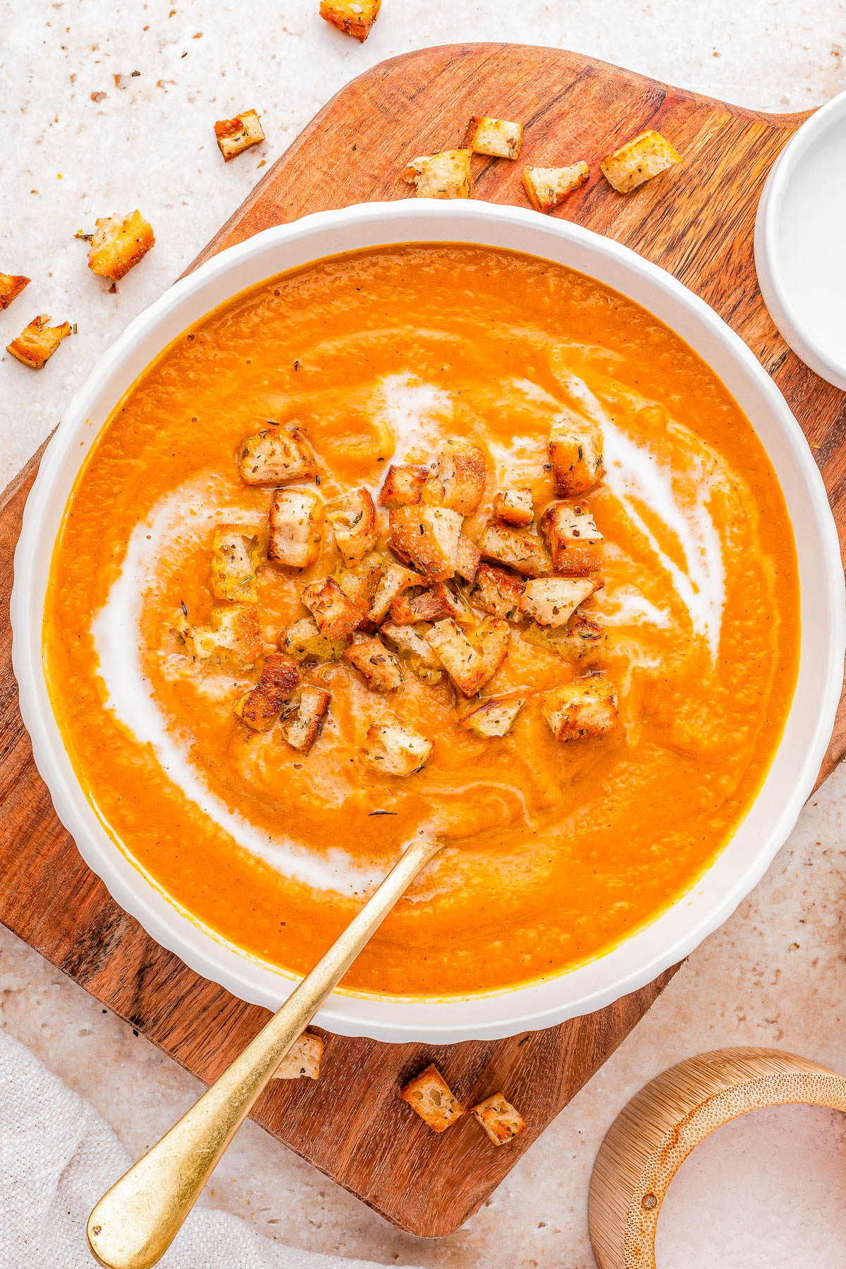 A bowl of creamy orange soup topped with croutons and a swirl of cream, placed on a wooden board with a gold spoon and scattered croutons nearby.