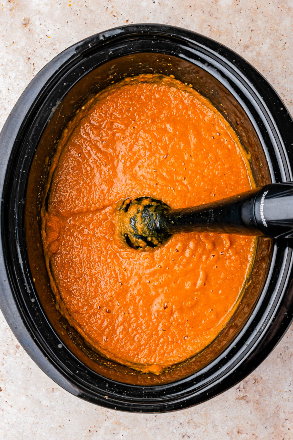 A black immersion blender is blending a thick orange sauce or soup inside a black slow cooker, viewed from above.