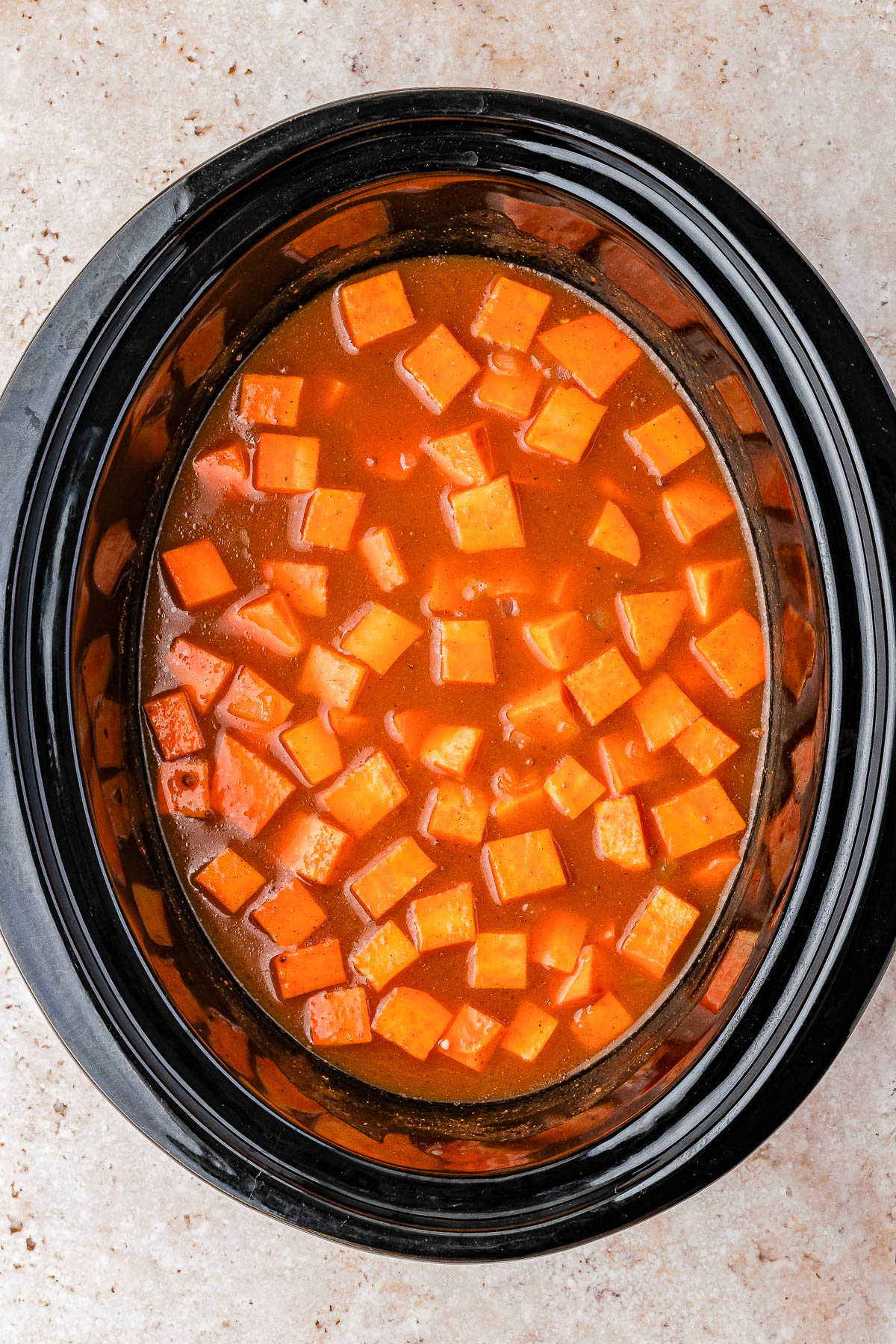 Cubed sweet potatoes in a red-orange sauce inside a black slow cooker on a light textured surface.