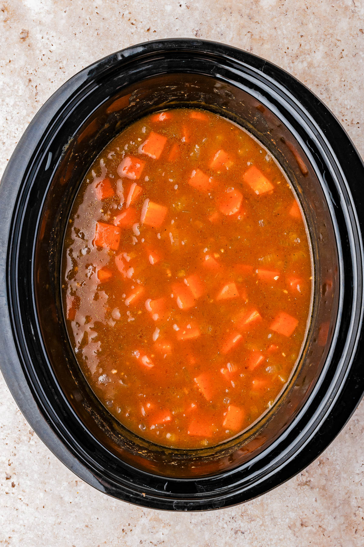 A slow cooker filled with chunky vegetable soup in a reddish broth, containing diced carrots and other vegetables, on a light-colored countertop.