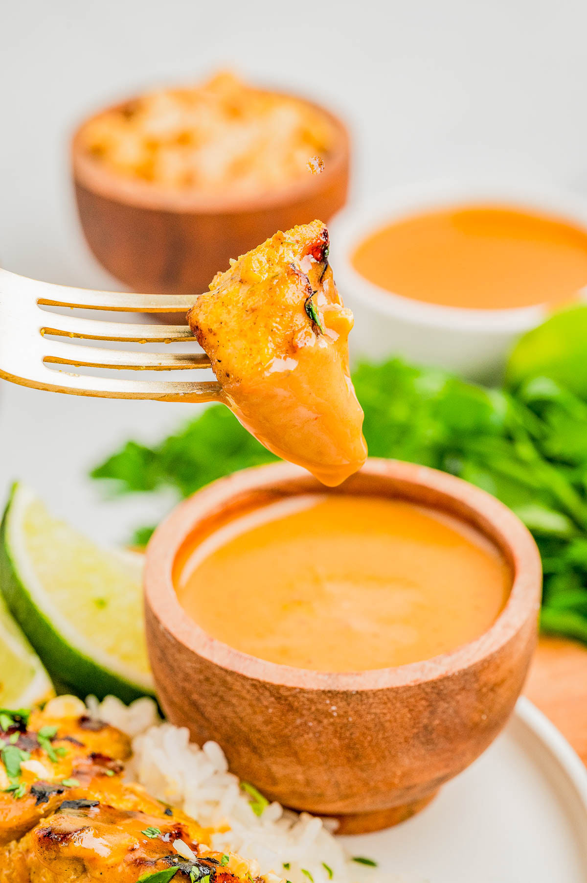 A fork holds a piece of grilled chicken above a small wooden bowl filled with orange dipping sauce; rice, lime, and greens are in the background.