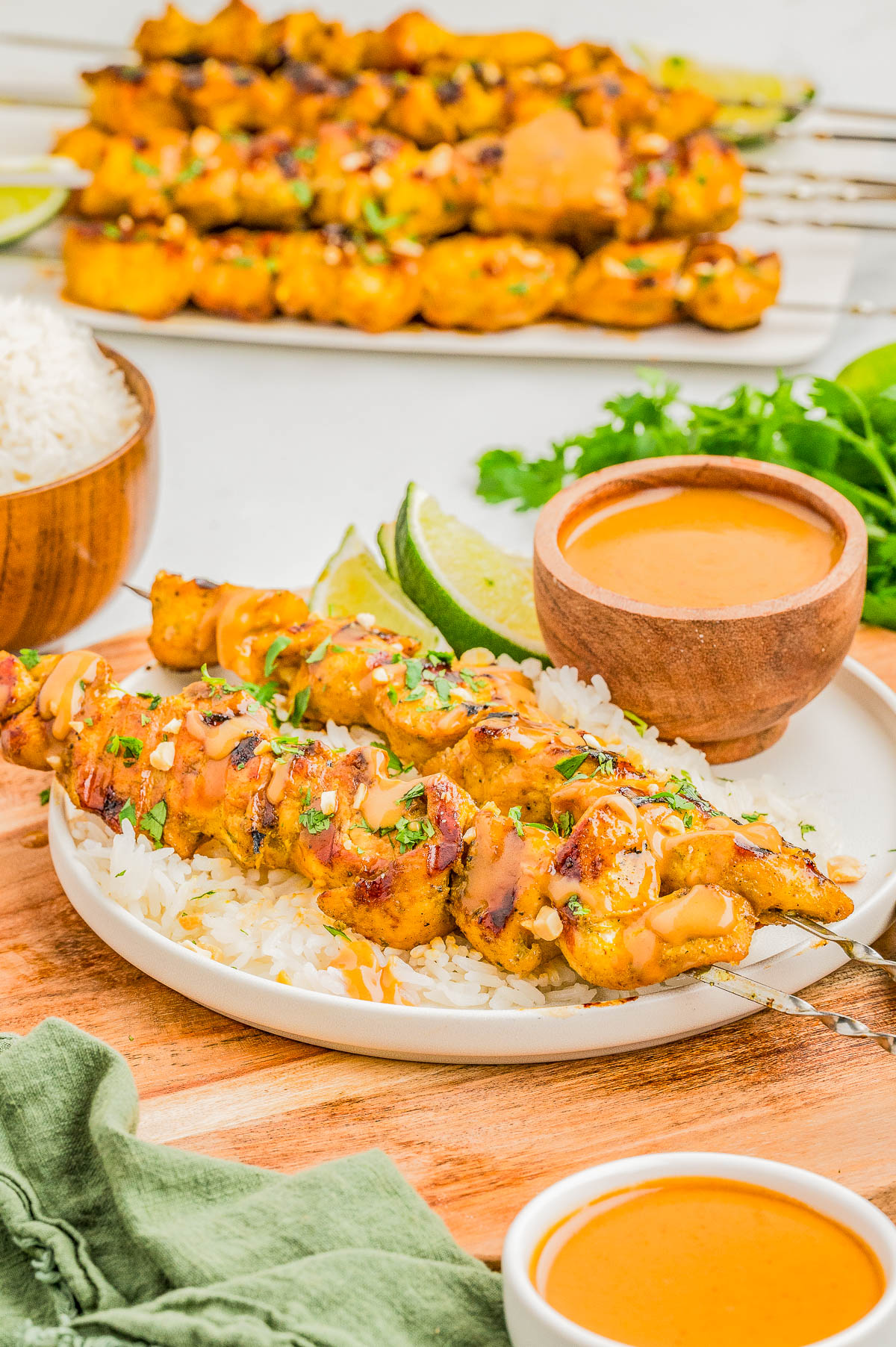 Chicken skewers on white rice, topped with a creamy orange sauce, served with lime wedges and herbs. Extra sauce, rice, and grilled skewers visible in the background.