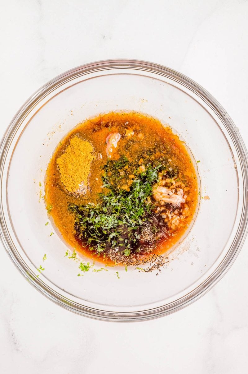A glass bowl containing oil, ground turmeric, herbs, ground pepper, and other seasonings on a white surface.