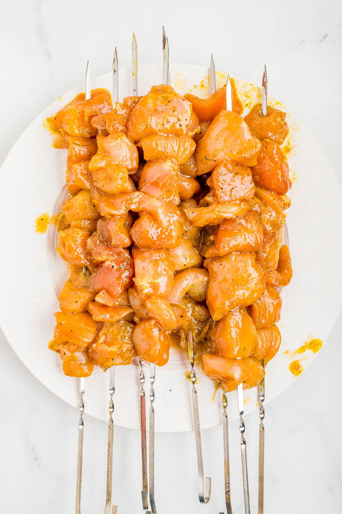 Raw marinated chicken pieces skewered on metal sticks, arranged on a white plate, ready for grilling or cooking.