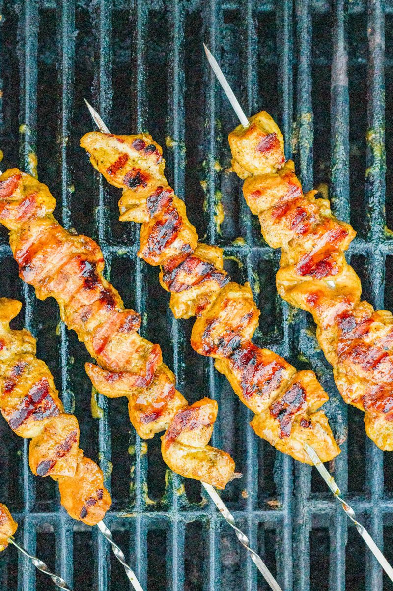 Four skewers of marinated chicken pieces grilling on a barbecue, with visible grill marks and charred edges.