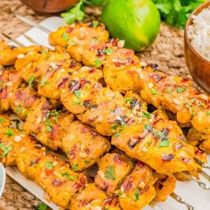 Grilled chicken skewers garnished with herbs and sauce, served on skewers with a lime, peanuts, rice, and fresh cilantro in the background.