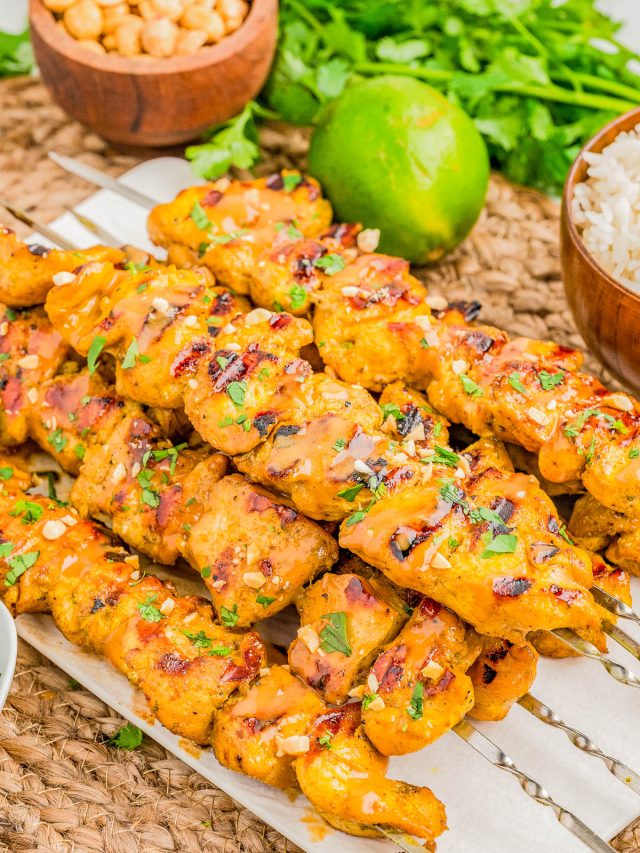 Grilled chicken skewers garnished with herbs and sauce, served on skewers with a lime, peanuts, rice, and fresh cilantro in the background.