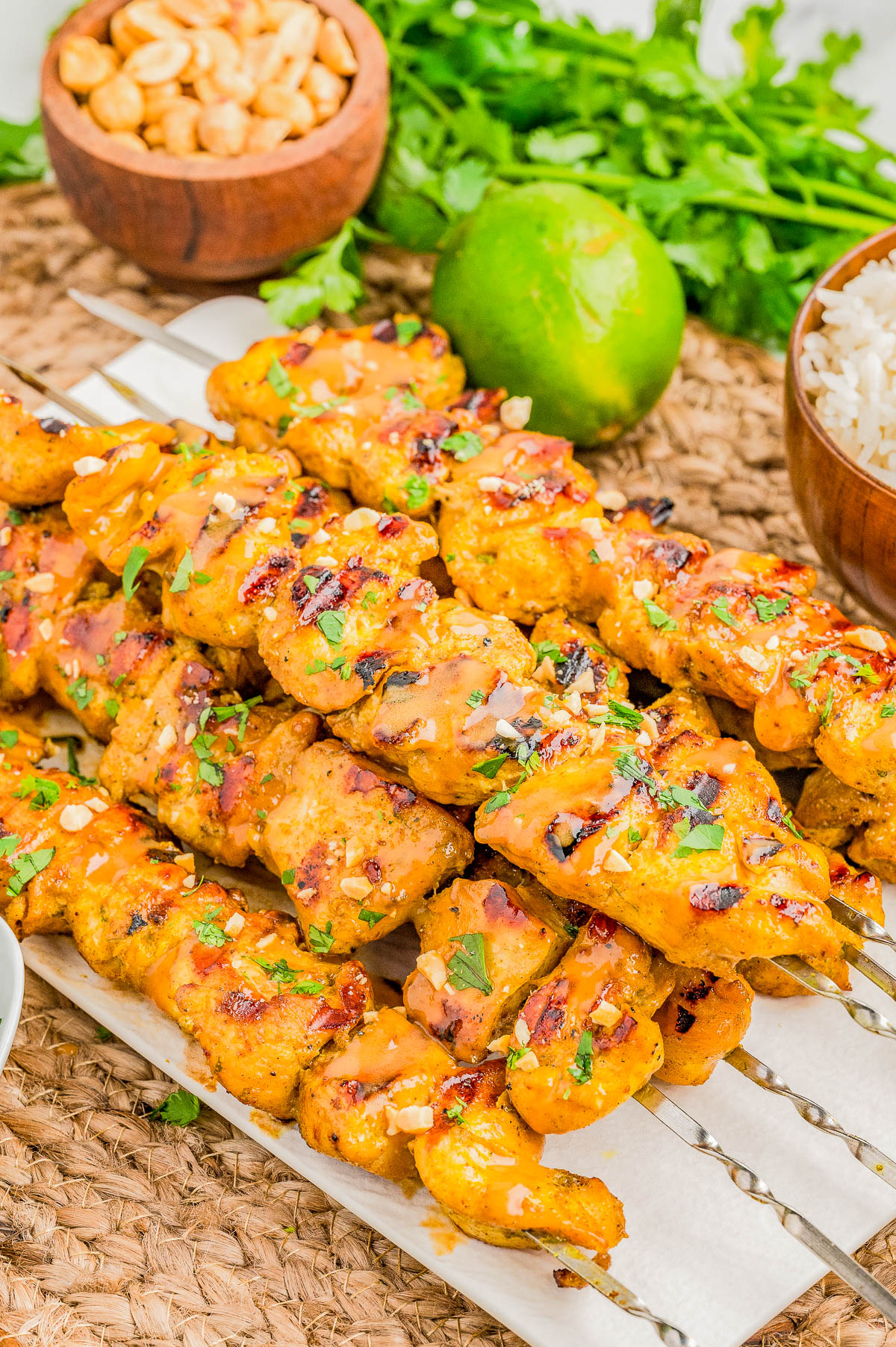 Grilled chicken skewers garnished with herbs and sauce, served on skewers with a lime, peanuts, rice, and fresh cilantro in the background.