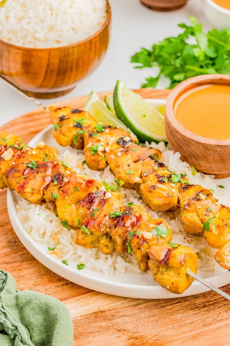 Grilled chicken skewers served on white rice with a side of dipping sauce, lime wedges, and garnished with chopped herbs.