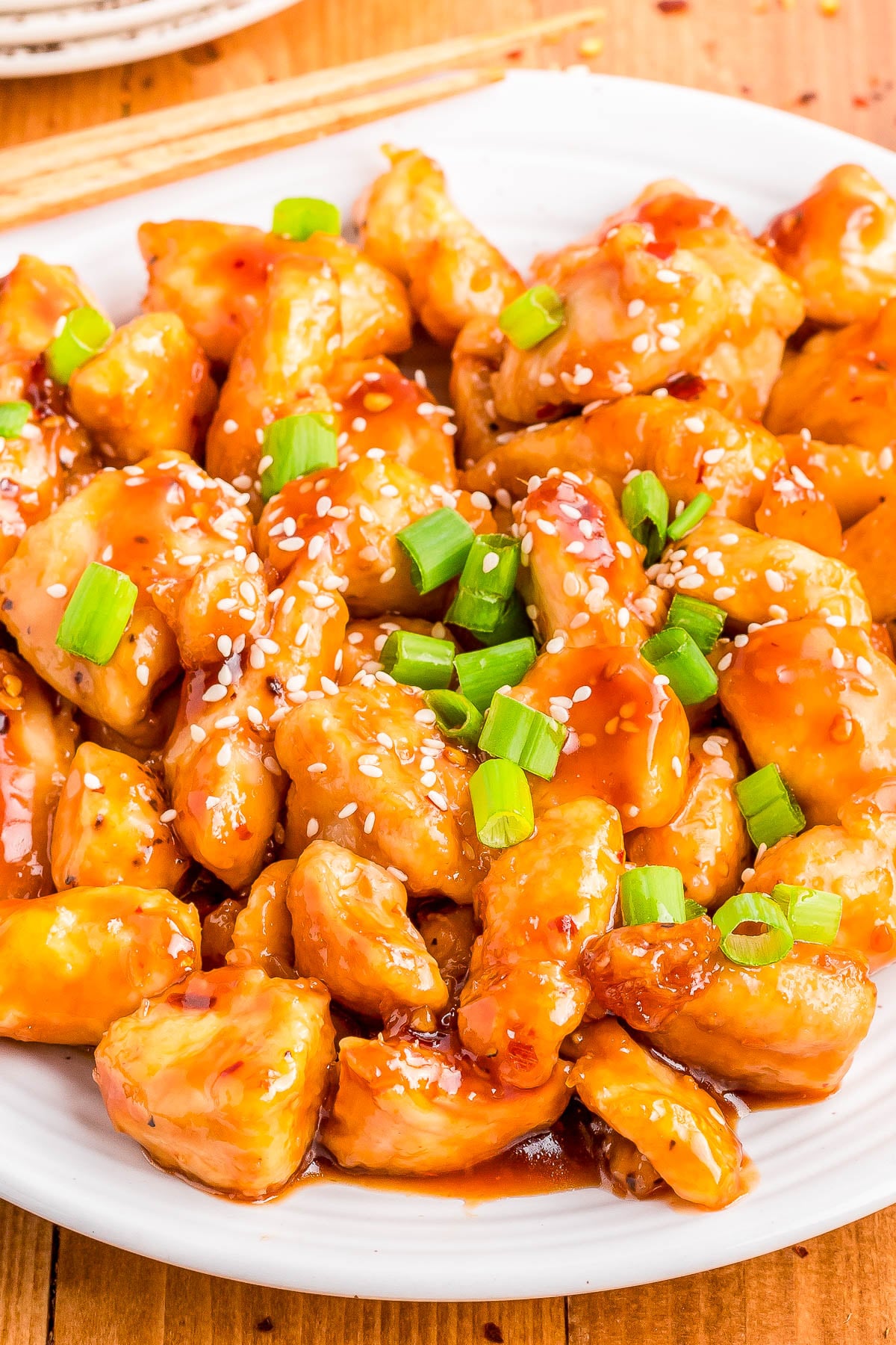 A plate of saucy chicken pieces garnished with chopped green onions and sesame seeds, with chopsticks visible in the background.