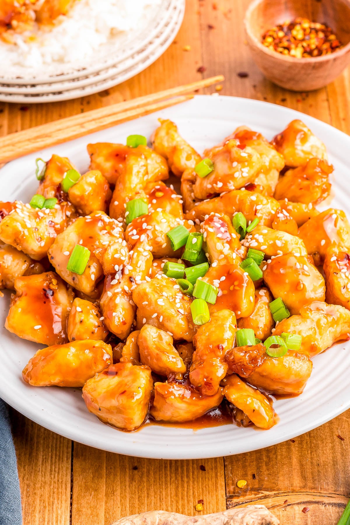 A plate of orange chicken topped with sesame seeds and chopped green onions, served on a white plate with chopsticks on a wooden table.