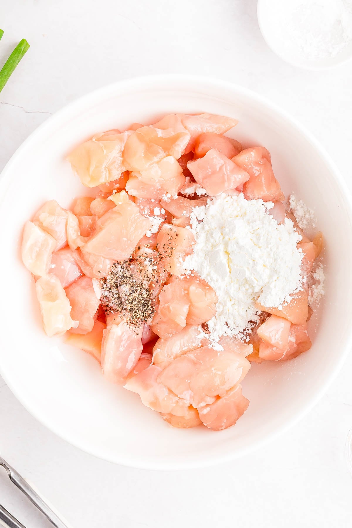 Raw diced chicken breast in a white bowl, topped with cornstarch, black pepper, and salt, on a light countertop.