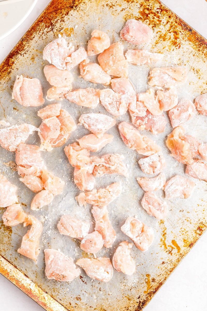 Raw, flour-coated chicken pieces spread out on a well-used baking sheet.