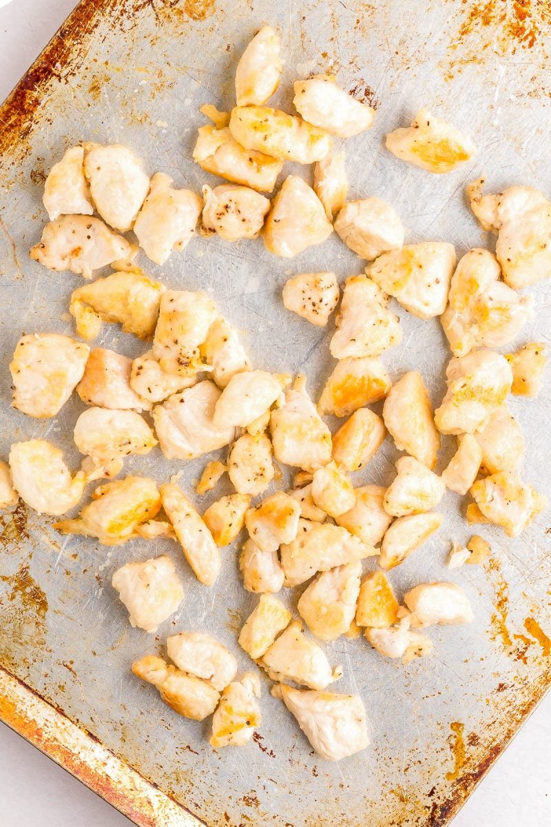 Cooked, diced chicken pieces are scattered on a well-used, lightly stained metal baking sheet.