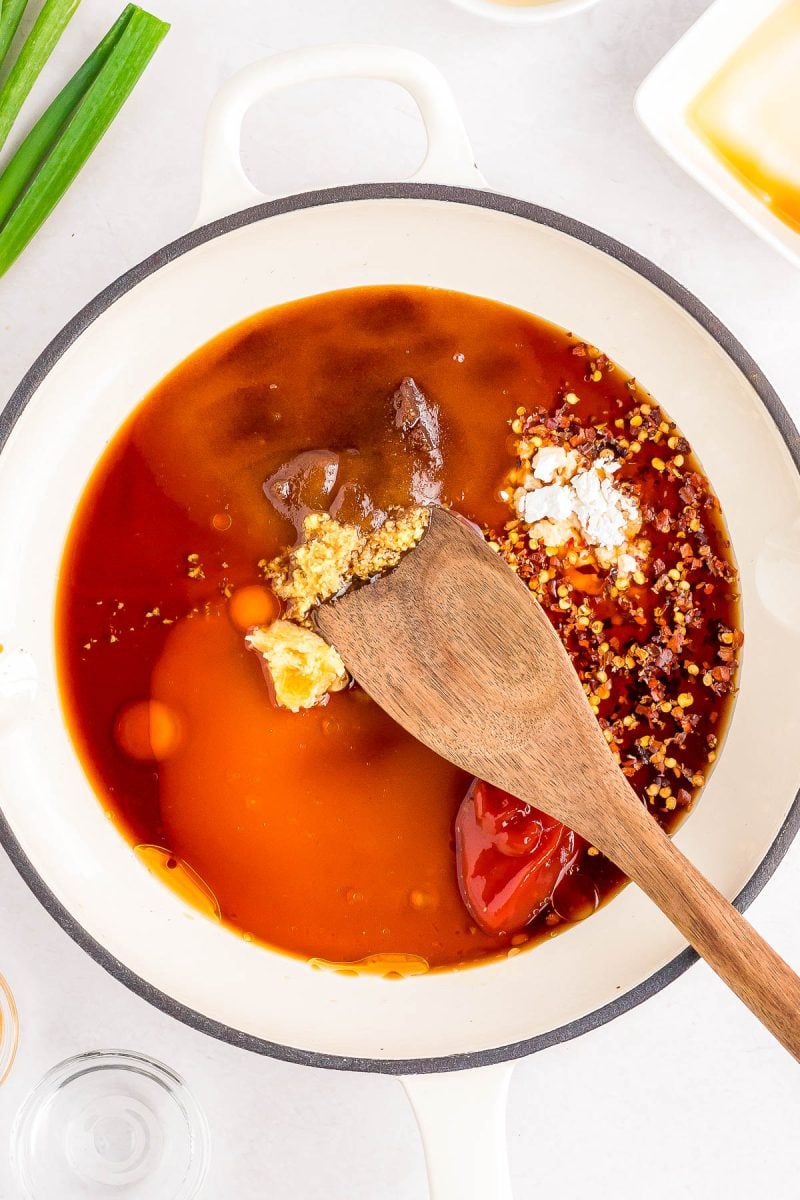 A wooden spoon rests in a white pot filled with various uncooked sauce ingredients, including brown paste, minced garlic, chili flakes, and soy sauce.