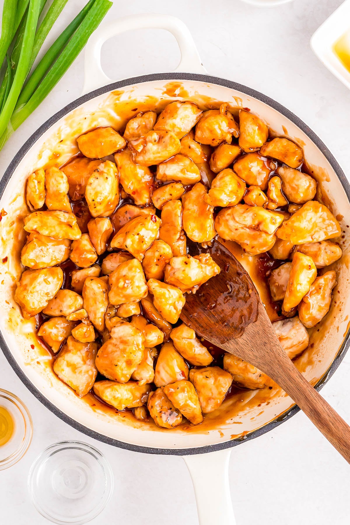 A white pan filled with glazed chicken pieces, stirred with a wooden spoon. Green onions and small bowls are nearby on a white surface.